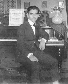 Un chico joven mira a cámara de espaldas a un piano en el que apoya el codo.