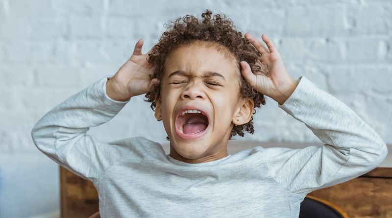 Despaired African American boy with curly hair and closed eyes screaming while standing in light room at home near wall