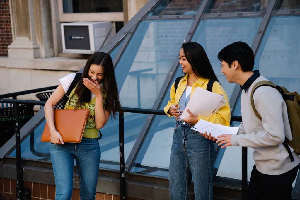 Three students sharing a laugh while standing outside a university building. Perfect for education and campus life themes.