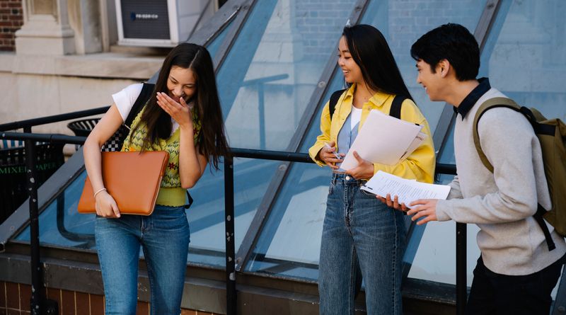 Three students sharing a laugh while standing outside a university building. Perfect for education and campus life themes.