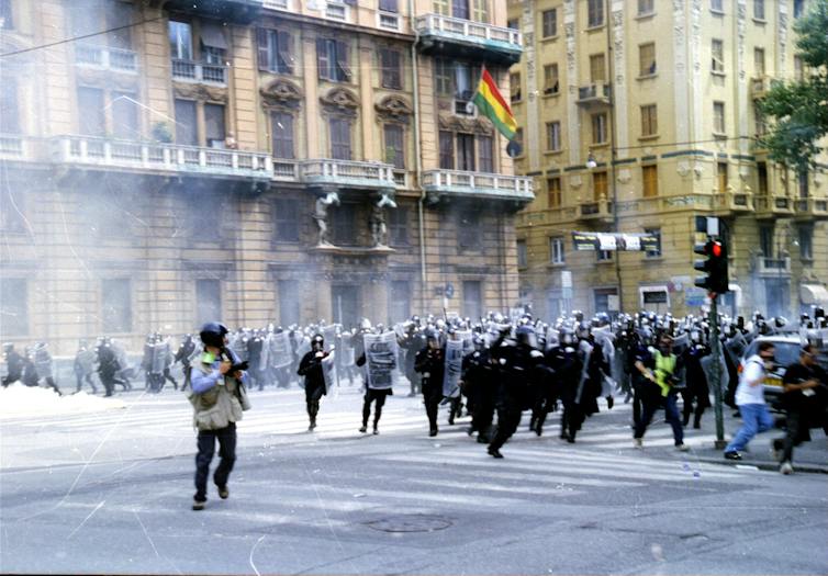 Imagen de fotógrafos de prensa corriendo delante de unos policías que cargan durante unas protestas en la calle de una ciudad.